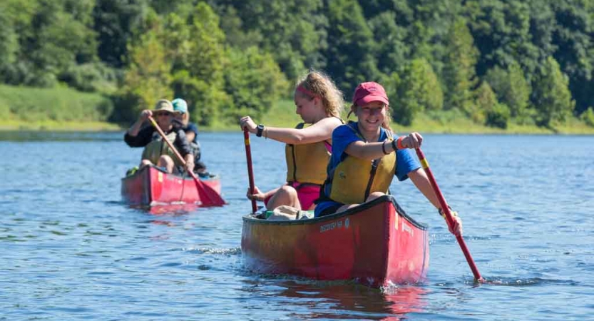 Two people each paddle two red canoes toward from the camera. They are wearing life jackets and there is a tree-lined shore in the background. 